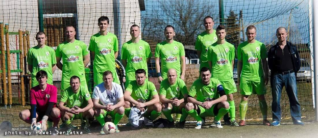 Eine Gruppe von Männern in grünen T-Shirts, einige in Hockstellung, steht auf einem Fußballfeld mit einem Ball und einem Tor, umgeben von Bäumen, Gebäuden und Fahrzeugen.