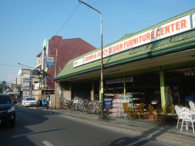 Eine belebte Straße mit fahrenden Fahrzeugen, parkenden Fahrrädern, Gebäuden mit Namensschildern, Laternenmasten und Strommasten mit Oberleitungen, mit einem Möbelsgeschäft im Vordergrund und einem sichtbaren Himmel im Hintergrund.