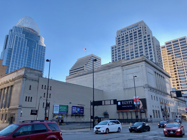 Eine belebte Stadtstraße in der Innenstadt von Cincinnati, Ohio, mit Autos, hohen Gebäuden, Laternenmasten, Ampeln, Texttafeln, einer Fahne an einem Mast und sichtbarem Himmel darüber.