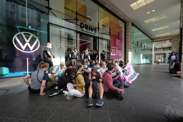 Menschen sitzen vor einem Volkswagen-Hndler auf dem Boden und halten eine Plakette waehrend einer Demonstration.