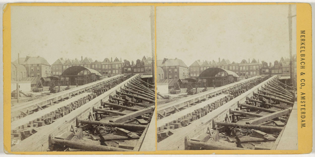 Schwarz-weiß-Foto einer Zugschiene mit Gebäuden im Hintergrund, Text 'Merkelbach & Co, Co. Amsterdam' rechts und verstreute hölzerne Gegenstände im Vordergrund.