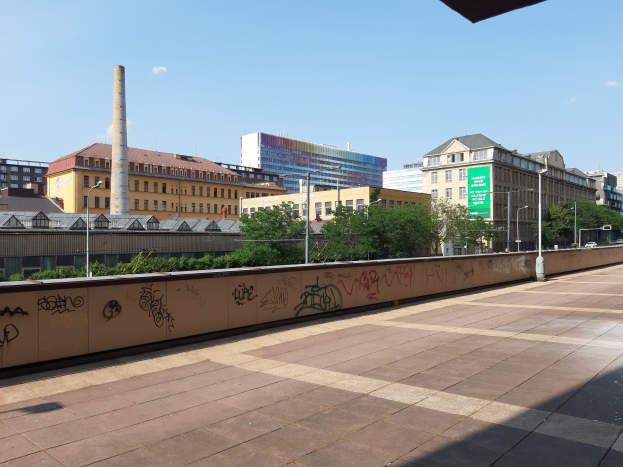 Stadtstraßenansicht mit Graffiti-bedeckter Wand, Bäumen, Straßenlaternen, Hinweisschild, Fahrzeugen, Gebäuden mit Fenstern, einem Schornstein und einem bewölkten Himmel.