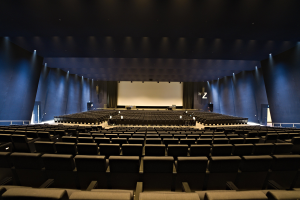 Ein großer Zuschauerraum mit Sitzreihen, Projektionsschirm und Bühnenobjekten, beleuchtet von Wandlampen.