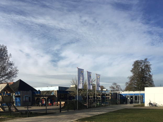 Großes Gebäude mit Metallzaun, Rasenfläche, Weg, Bäumen, Schildern, Fahrrad, nahen Häusern und bewölktem Himmel, als neuer Spielbereich für Kinder an der Schule identifiziert.