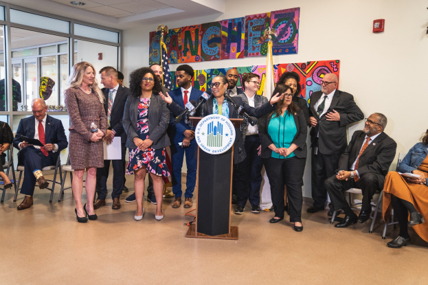 Gruppe von Menschen vor einem Podium mit Mikrofon und einer Tafel mit der Aufschrift "Sancho-Stadtrat tritt dem Stadtrat bei", mit sitzenden und stehenden Teilnehmern, Flaggen, Feuerlöscher und Deckenbeleuchtung.