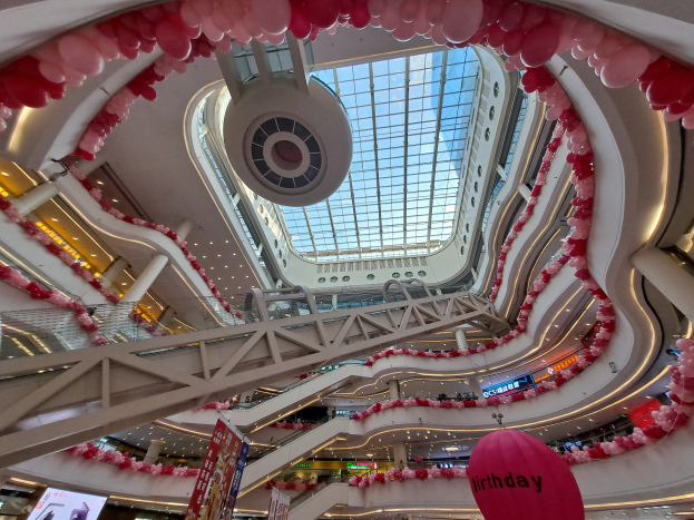 Ein Einkaufszentrum von innen mit pinken und weißen Ballons geschmückt, Rolltreppen, Geländer, Texttafeln, Lichter und eine Glasdecke.