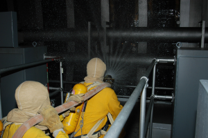 Zwei Feuerwehrleute in gelben Uniformen, Helmen und Handschuhen sprühen Wasser aus einem Schlauch auf ein Metallgeländer, mit Metallstangen und anderen Gegenständen im Hintergrund und Text unten.