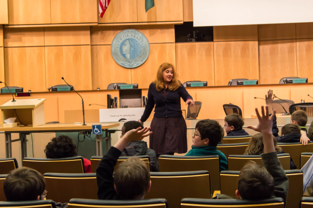 Eine Frau, die als die Seattle Superintendent identifiziert wurde, steht an einem Podium mit Mikrofonen in einem Klassenzimmer voller aufmerksamer Kinder, mit einem Logo und zwei Flaggen an der Wand dahinter.