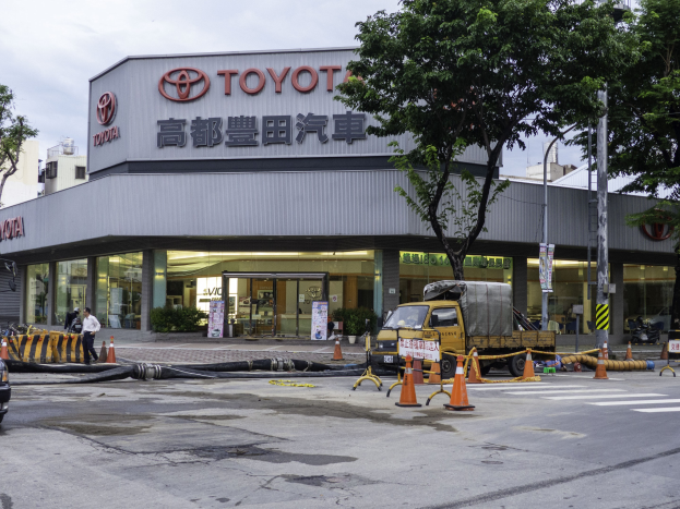Außenansicht einer Toyota-Niederlassung in Tokio, Japan, mit Fahrzeugen, Verkehrskegeln, Rohren, Fußgängern, Bäumen, Strommasten, Schildern und umliegenden Gebäuden unter einem bewölkten Himmel.