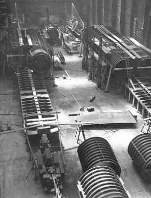 Schwarzes und weißes Foto eines industriellen Fabrikinneren mit ausgedehnten Rohrleitungen, Maschinen und Geräten auf dem Boden, vor strukturellen Säulen und einer Wand.