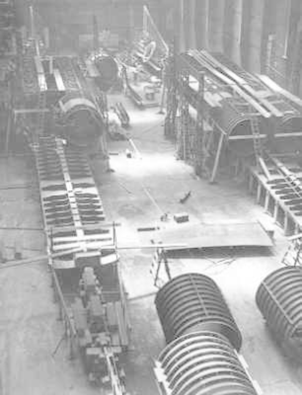 Schwarzes und weißes Foto eines industriellen Fabrikinneren mit ausgedehnten Rohrleitungen, Maschinen und Geräten auf dem Boden, vor strukturellen Säulen und einer Wand.
