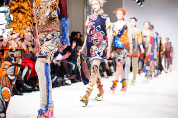 Eine Gruppe Von Frauen, Die Auf Einem Laufsteg In London Fashion Week Gehen, Mit Sitzenden Zuschauern Und Heller Beleuchtung Im Hintergrund, Die Szenerie Etwas Unscharf.