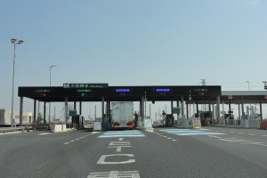 Eine Mautstelle mit einem vor ihr parkenden Lastwagen, neben einer mit wei├č-blauen Linien markierten Stra├če, Lichtmasten, Schildern und anderen Gegenst├Ąnden, mit Geb├Ąuden, B├Ąumen und einem klaren Himmel im Hintergrund.