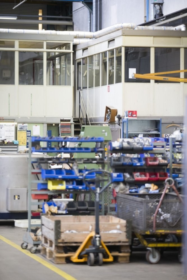 Großer Lagerraum mit verschiedenen Geräten einschließlich fahrbaren Wagen, Schränken und einer Box, mit einer Glasfensterwand und freiliegenden Rohren im Hintergrund.