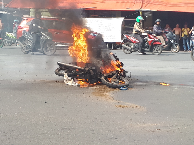 Motorrad in Flammen auf der Straße mit Menschen in der Nähe, einem Auto und einem Van im Hintergrund, einer Fahne und einem Zelt.