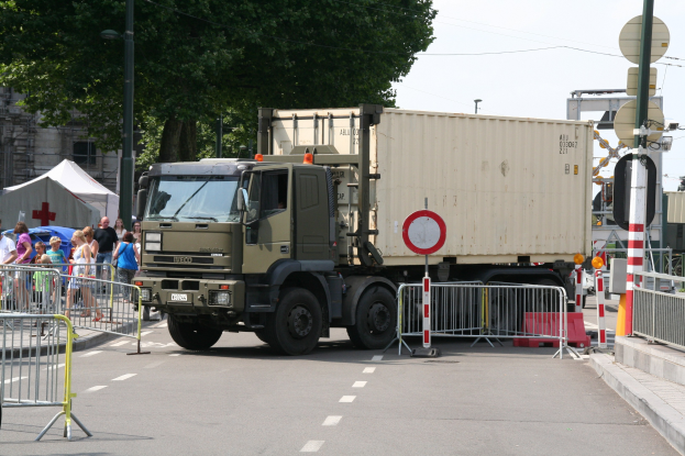 Ein Lkw am Straßenrand mit einer Gruppe von Menschen auf dem Gehweg, umgeben von Absperrungen, Schildern, Polen, Lampen, Kabeln, Bäumen, Gebäuden und einem klaren blauen Himmel.