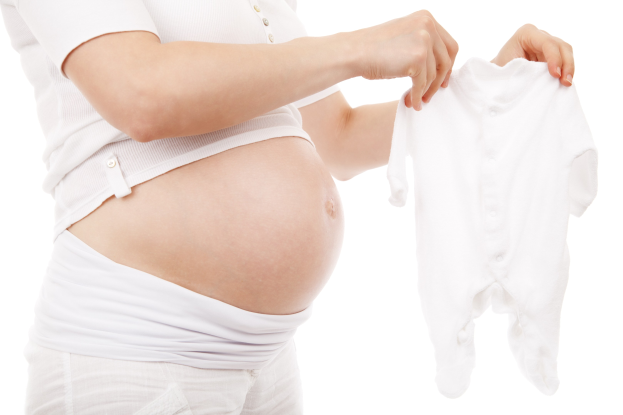 Eine schwangere Frau in einem weißen Kleid hält ein weißes Baby- Onesie und strahlt glücklich.