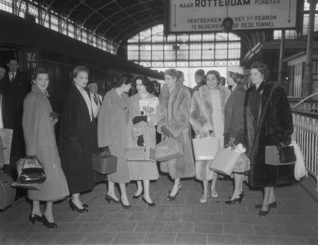 Schwarzes und weißes Bild einer Gruppe von Frauen, die an einem Bahnhof stehen und Taschen halten, mit einem Zug links und Geländern und einer Tafel rechts.