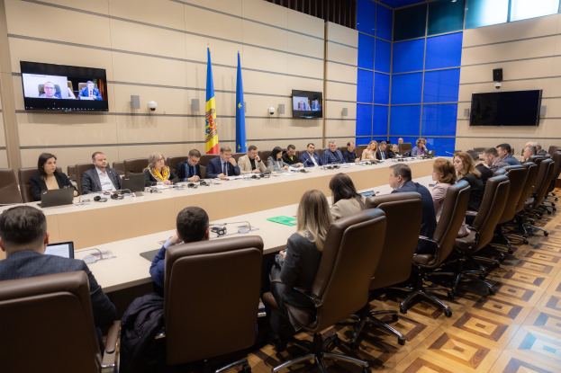 Eine große Gruppe von Menschen sitzt um einen langen Tisch mit Laptops, Mikrofonen, Papieren und anderen Gegenständen während einer Sitzung des Europäischen Rates für Menschenrechte, mit drei Flaggen, zwei Fernsehern und Deckenleuchten im Hintergrund.