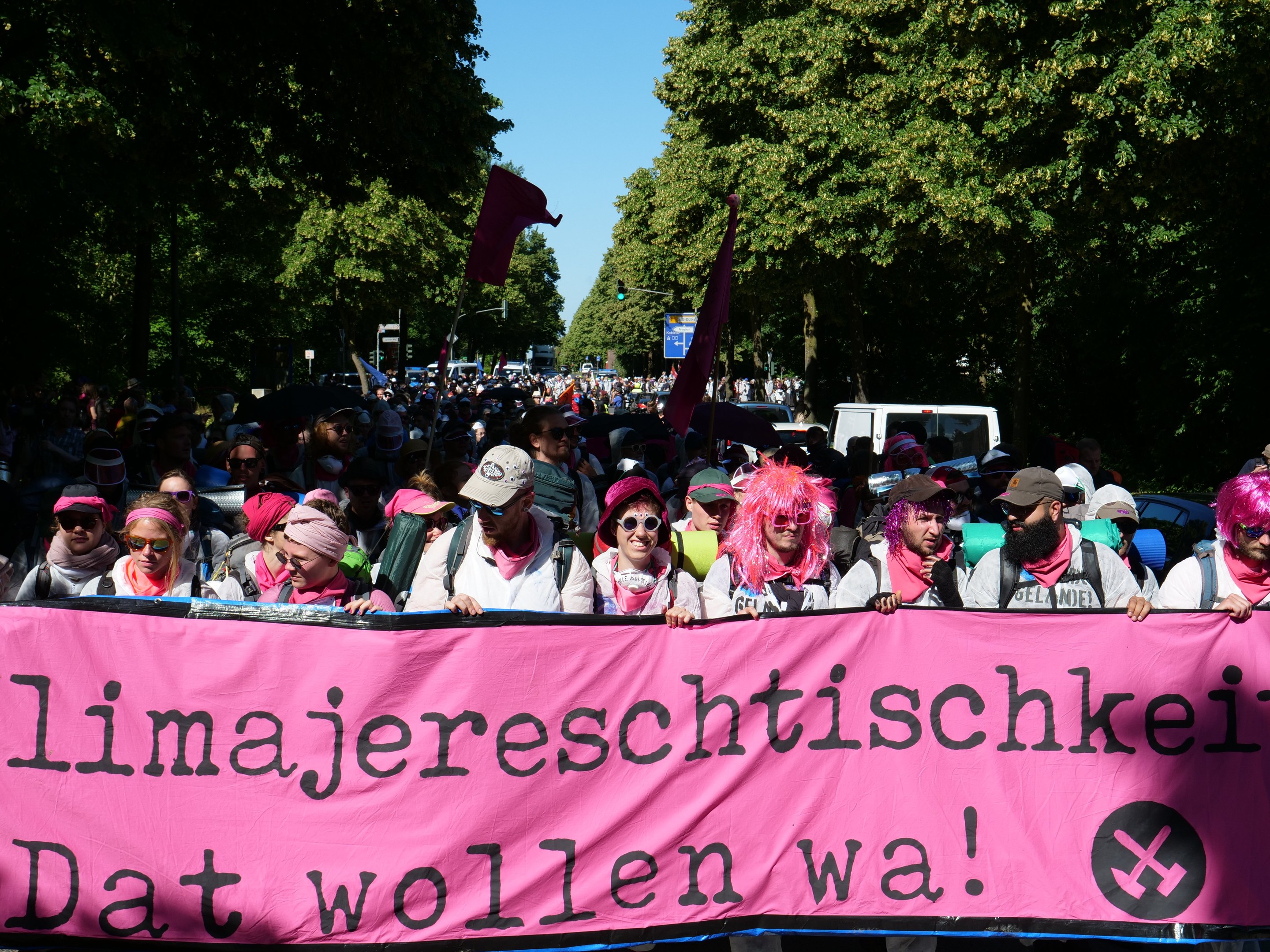 Eine Gruppe von Menschen marschiert die Straße entlang, hölt eine pinkfarbene Banner mit der Aufschrift "Women's March in Berlin", mit einigen, die Köpfe und Schutzbrillen tragen und andere Fahnen halten, vor dem Hintergrund von Bäumen, Fahrzeugen und Himmel.