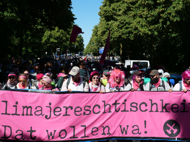 Eine Gruppe von Menschen marschiert die Straße entlang, hölt eine pinkfarbene Banner mit der Aufschrift "Women's March in Berlin", mit einigen, die Köpfe und Schutzbrillen tragen und andere Fahnen halten, vor dem Hintergrund von Bäumen, Fahrzeugen und Himmel.