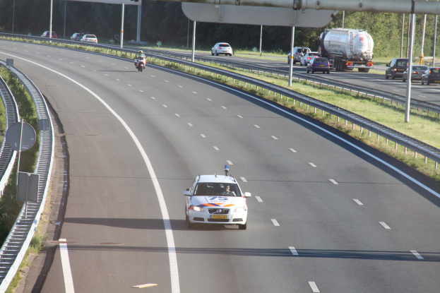 Ein Auto auf einer Autobahn mit einem stationären Polizeiwagen am Röstrand, andere Fahrzeuge und Bäume, Geländer, Masten und Planken im Hintergrund.