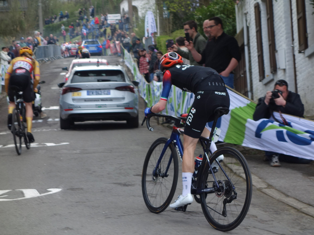 Eine Gruppe von Radfahrern fährt auf einer Straße mit Autos nebenan, Menschen mit Kameras und einem Banner auf der rechten Seite und im Hintergrund Gebäude, Bäume, ein Zaun und Himmel.