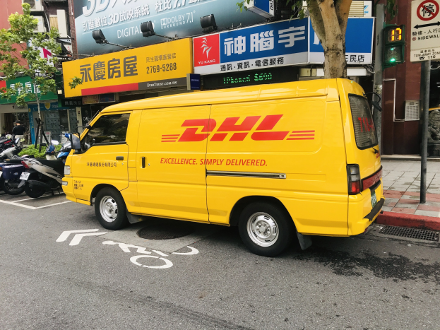 Gelber DHL-Lieferwagen auf der Straßenseite geparkt, umgeben von Gebäuden, Bäumen, Schildern, Pfählen, Verkehrssignalen und einer Person auf dem Gehweg.