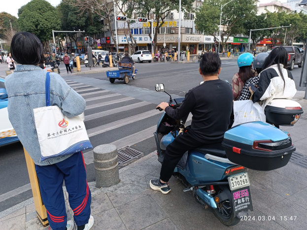 Eine Gruppe von Menschen auf dem Rücksitz eines blauen Roller, mit einer Frau auf dem Gehweg links daneben und verschiedenen Fahrzeugen, Fußgängern und Gebäuden im Hintergrund.