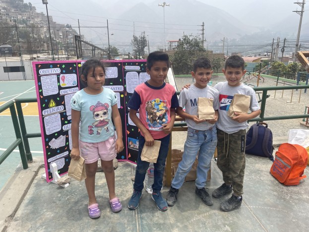Gruppe von Kindern auf einem Basketballfeld mit Papiertüten in den Händen, mit Gegenständen auf dem Boden rechts und einer Tafel mit Text links, vor dem Hintergrund von Zäunen, Pfählen, Bäumen, Gebäuden, Hügeln und Himmel.