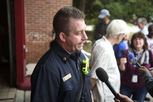 Ein Polizist spricht zu einer Gruppe von Menschen mit Mikrofonen, mit Bäumen und einem Gebäude im Hintergrund.