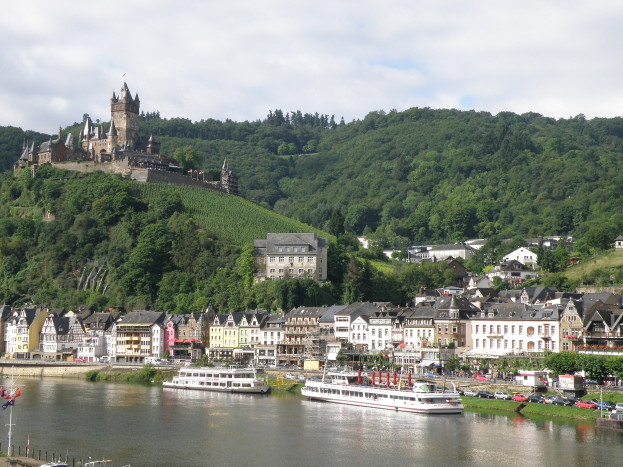 Ein idyllischer Blick auf den Rhein in Deutschland mit einer Burg auf einem Hügel, Booten auf dem Fluss, Fahrzeugen auf einer nahen Straße und einer bewölkten Himmel.