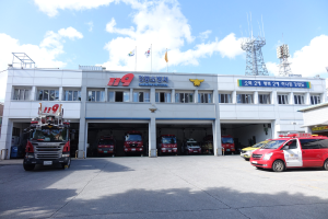 Feuerwache mit mehreren Feuerwehrautos vor dem Gebäude, Gebäude mit Fenstern und Text, Flaggen mit Stangen im Hintergrund, bewölkter Himmel und ferne Turm.