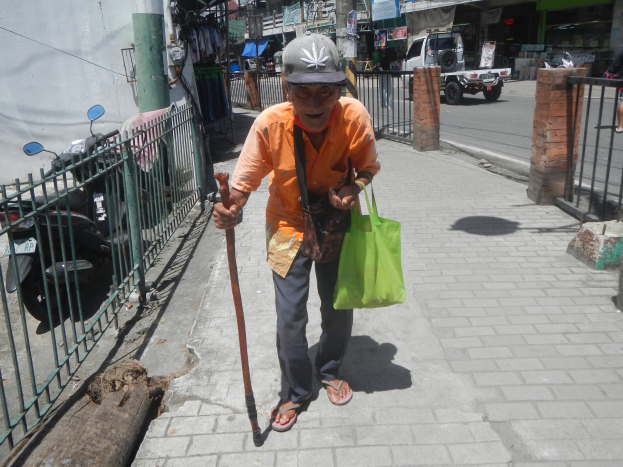 Ein älterer Mann geht die Straße entlang, in einer Hand einen Stock, in der anderen eine Tasche und einen Stock, trägt eine Mütze und ist von einem Metallzaun auf der linken Seite umgeben.