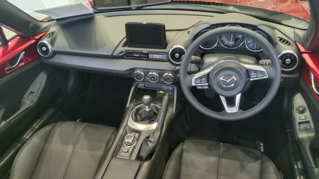 Innenansicht eines roten Mazda MX-5 Miata RF mit schwarzen Ledersitzen, Lenkrad, Schaltknüppel, Knöpfen, Bildschirm und Spiegel.