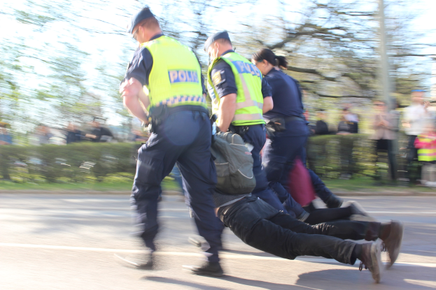 Eine Gruppe von Polizisten in Mützen schiebt einen Mann mit einer Tasche eine Straße entlang, umgeben von Bäumen, Pflanzen und einem Pfahl.