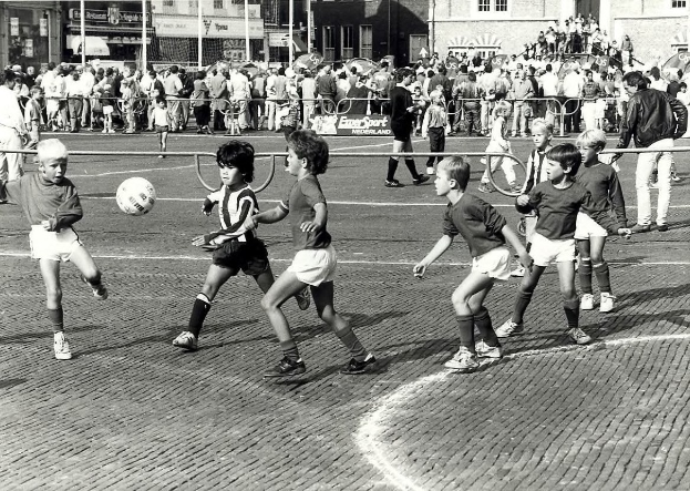 Schwarzes Foto von jungen Jungs, die Fussball auf der Straße spielen, umgeben von einer Zuschauermenge hinter einem Geländer, Gebäuden und Texttafeln im Hintergrund.