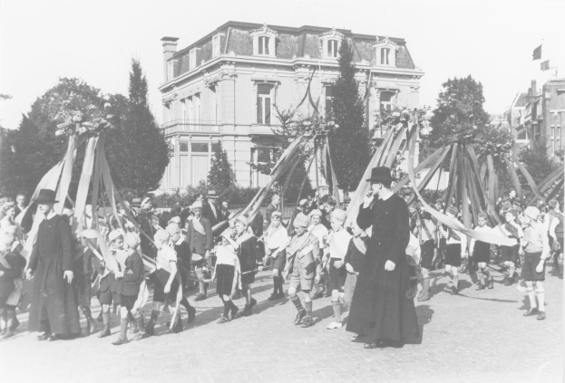 Ein Schwarz-Weiäs Bild einer Gruppe von Menschen, die eine Straäe entlanggehen, einige halten Fahnen, mit Häuten, Bäumen, Gebäuden und einem sichtbaren Himmel im Hintergrund, das einen Maypole zeigt, der auf ein May Day-Fest hinweist.