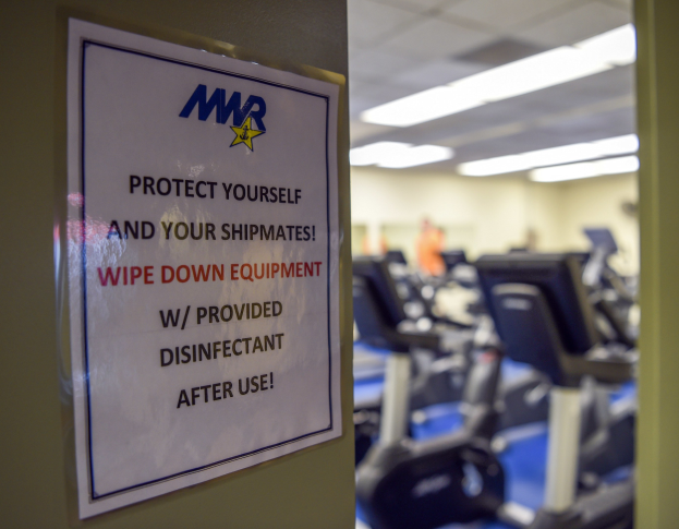 Ein Türschild mit der Aufschrift "Schützen Sie sich und Ihre Schiffskameraden" mit Laufbändern und Deckenlampen im Hintergrund, mit einem leicht unscharfen, bewegungsähnlichen Effekt.