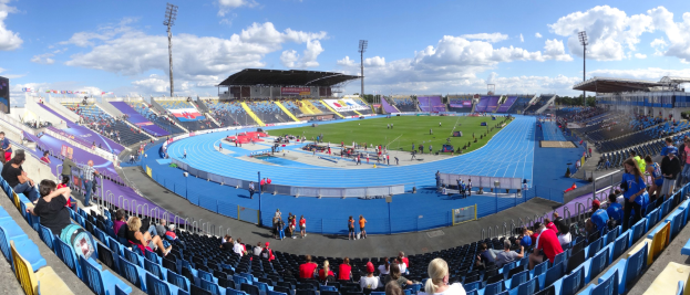 Großes Stadion voller Zuschauer bei einer Leichtathletikveranstaltung, mit Sitzplätzen und Stehplätzen, Flutlicht, Hütten, Bäumen und einem bewölkten Himmel im Hintergrund.