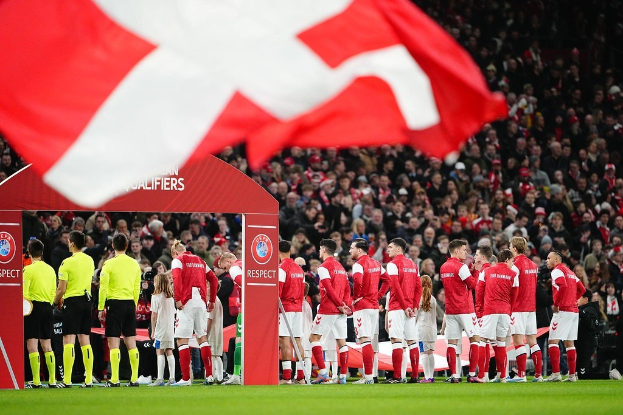 Eine Gruppe von Menschen auf einem Fußballfeld mit einer roten und weißen Flagge im Vordergrund, einem Bogen mit der Aufschrift "Bayern München vs. Bayern München Wetten & Vorschau" im Hintergrund und einer großen Menschenmenge im Stadion.