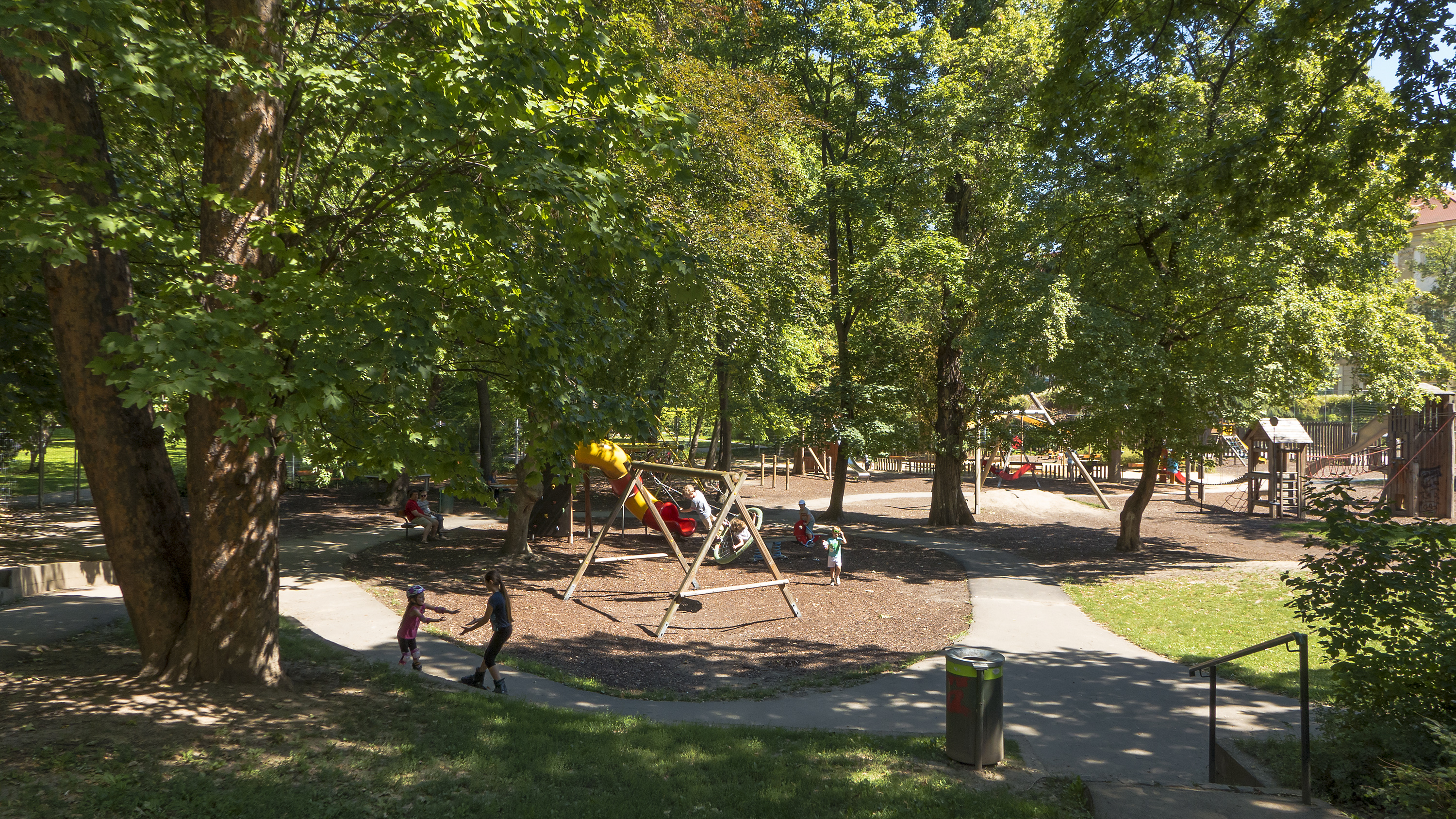 Ein Park mit einem Spielplatz in der Mitte, umgeben von Bäumen, mit Kindern auf der Ausrüstung, einem Mülleimer, Geländern, Pflanzen, Gras und Gebäuden im Hintergrund unter einem klaren blauen Himmel.