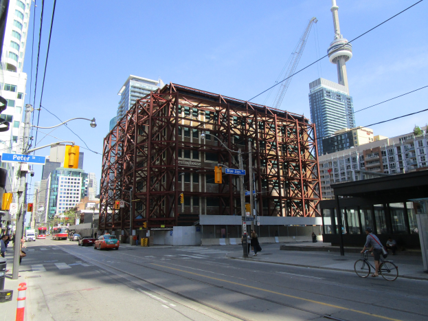 Eine belebte Stadtstraße mit einem großen Baugerüst im Hintergrund, Fahrzeuge auf der Straße, ein Radfahrer, Fußgänger auf dem Gehweg, Verkehrsampeln, Pfosten mit Schildern, ein Kran und der Himmel.