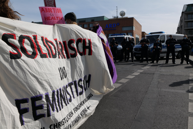 Gruppe von Menschen, die eine Straße entlanggehen und ein Banner mit der Aufschrift "Solidarität und Feminismus" tragen, mit parkenden Fahrzeugen und Gebäuden im Hintergrund unter einem klaren blauen Himmel.
