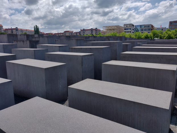 Konkreter Holocaust-Mahnmal mit rechteckigen Blöcken, die in einem Gittermuster angeordnet sind und von Bäumen, Gebäuden und einer bewölkten Landschaft umgeben sind.