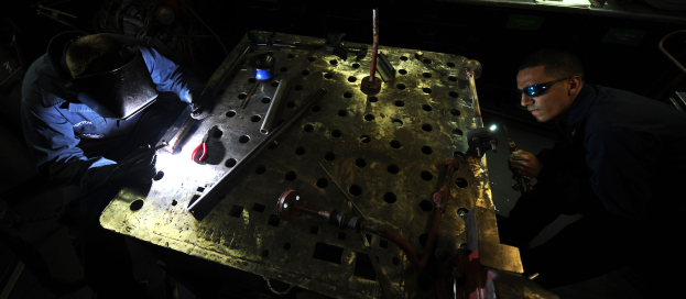 Zwei Männer in Schutzausrüstung arbeiten an einem Metallstück in einer schlecht beleuchteten Werkstatt, umgeben von verschiedenen Werkzeugen und Materialien auf einem Tisch.