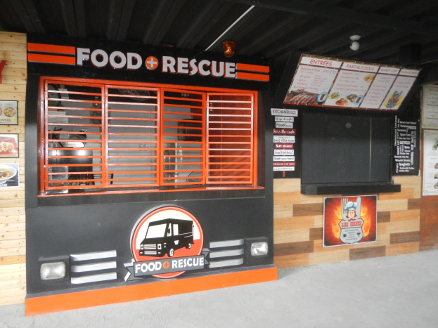 Ein Foodtruck vor einem Gebäude mit einer "Food Rescue"-Tafel, mit Fenster, Tür und Wandtafeln mit Text und Bildern.