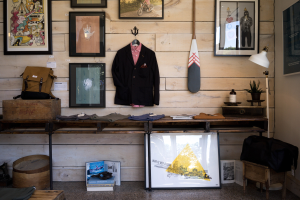 Ein unordentlicher Raum mit Holzverkleidung, die gerahmte Fotos, einen Mantel, Kleidung, eine Tasche, eine Schachtel, eine Flasche, eine Topfpflanze, eine Lampe und andere Gegenstände auf Tischen zeigt, mit einer Tafel mit Text auf der rechten Seite und einem Holzbehälter mit Gegenständen und einem Tuch auf einem Hocker auf der linken Seite, Teil der New York Fashion Week.