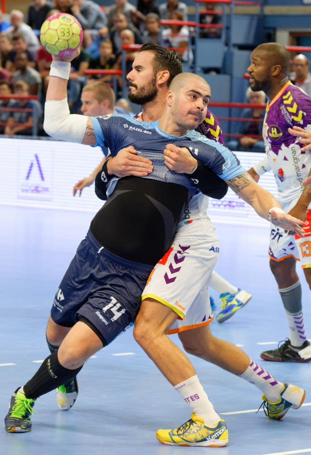 Männer spielen Hallenhandball, einer hält den Ball, mit Zuschauern auf Stühlen, Geländern und einer Wand in einer Halle.