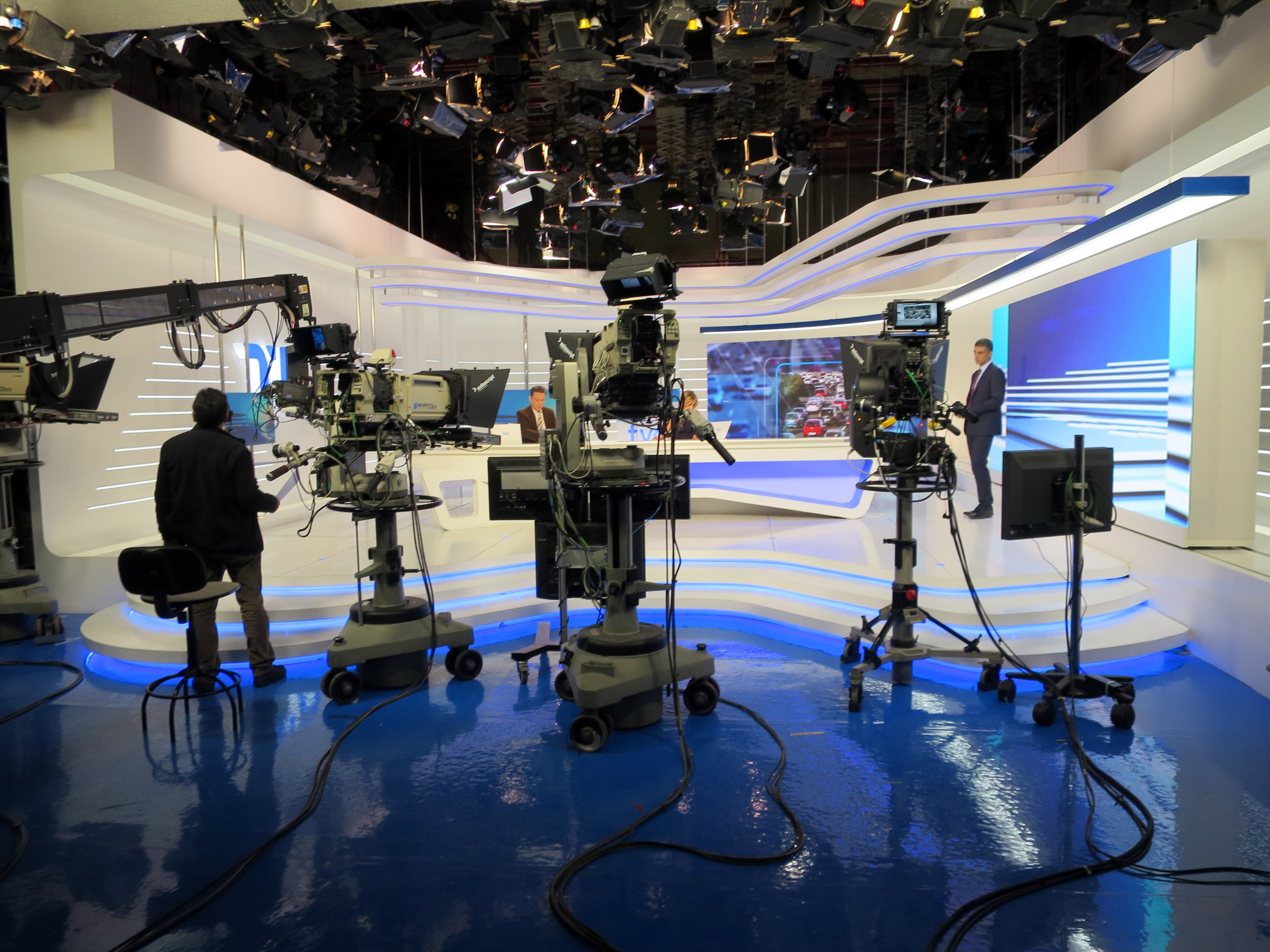 Ein TV-Studio mit mehreren Kameras auf dem Boden, einer sitzenden Person im Vordergrund und einigen stehenden Personen im Hintergrund, mit Deckenleuchten für eine Livesendung.